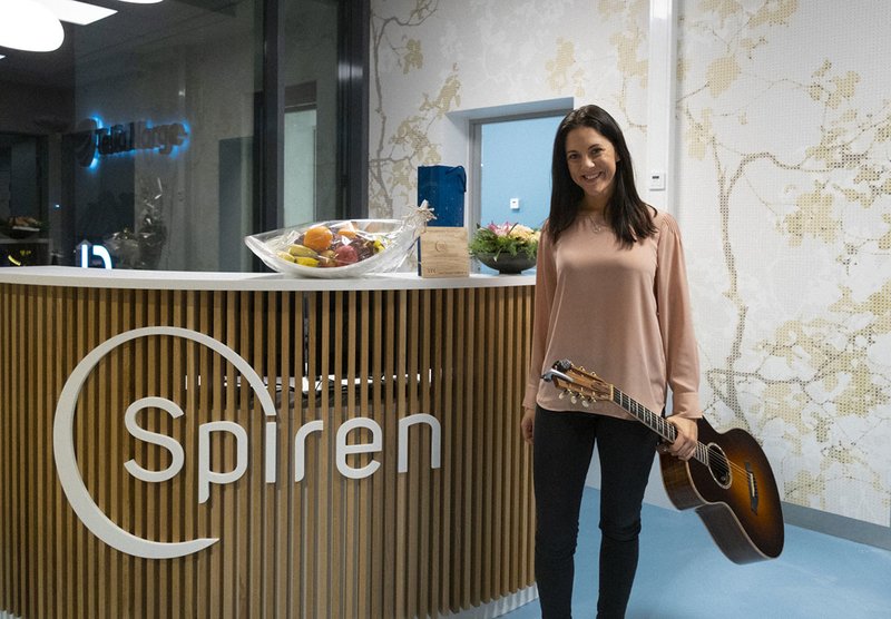 Ansatt i Spiren Fertilitetsklinikk som står stående smilende med en gavepakke med frukt og en gitar i hånden sin